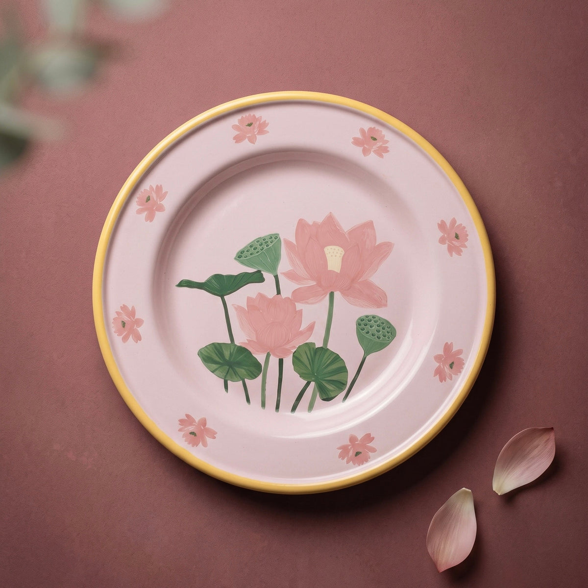 A round enamel ceramic plate with a pink background, yellow rim, and hand-painted lotus flowers and green leaves, styled on a dusty rose background with decorative petals.