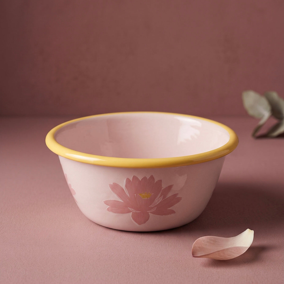 A light pink enamel bowl with a yellow rim and a pink lotus flower design, set against a matching dusty rose background with a single flower petal in the foreground.