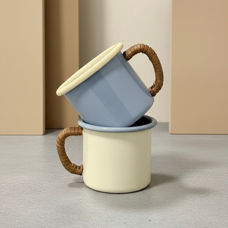 Stack of two enamel coffee mugs with leather wrapped handles on a neutral background
